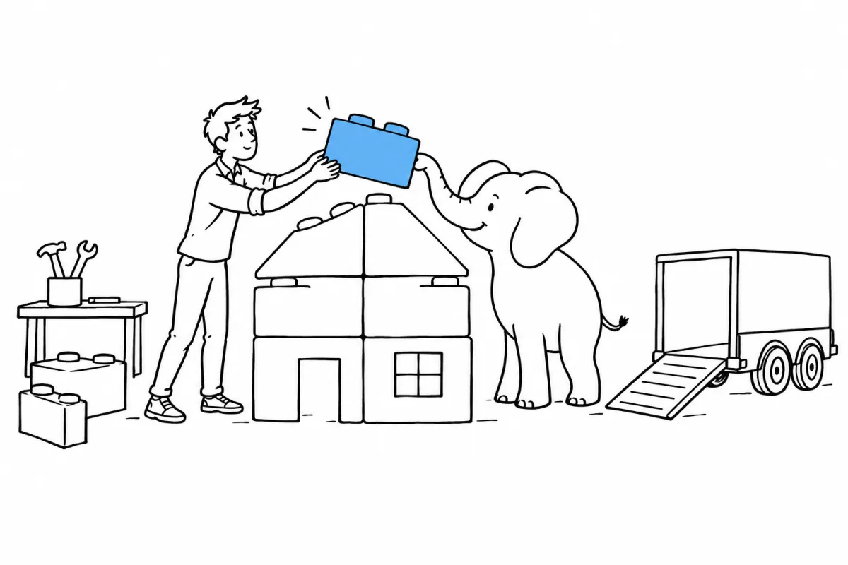 A person and an elephant place the last block, the only blue one, on a small house made of big building bricks. A trailer waits with its ramp down.